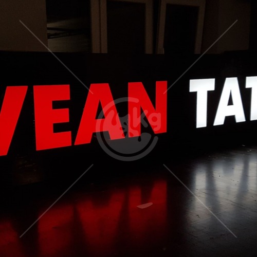 Częściowo widoczny napis 'VEAN TATTOO' w kolorach czerwonym i białym, wykonany z podświetlanych liter przestrzennych na czarnym tle, z odbiciem na błyszczącej powierzchni.