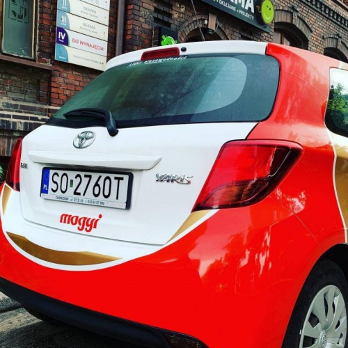 Toyota Yaris z oklejeniem reklamowym w kolorach biało-czerwono-złotym, z widocznym logo firmy 'moggi' na tylnej klapie, zaparkowana przed budynkiem z szyldami firm.
