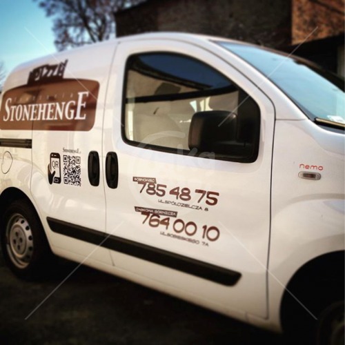 Biały samochód dostawczy z brązowym logo Stonehenge, kodem QR i numerami telefonów na karoserii, zaparkowany na zewnątrz budynku.