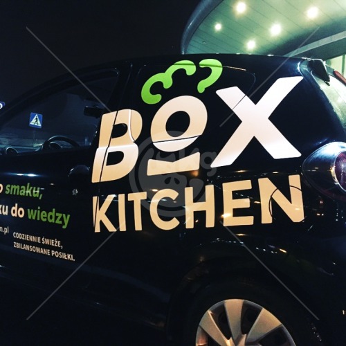 Czarny samochód z reklamą 'Box Kitchen' w Czeladzi, z białymi napisami i zielonym logo, zaparkowany na tle budynku.