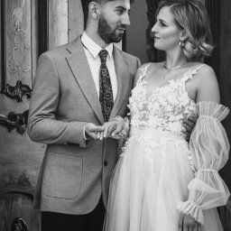 ASM PHOTOGRAPHY - Fotograf - Czarno-białe zdjęcie pary młodej, on w garniturze, ona w sukni ślubnej z koronką, trzymają się za ręce na tle rzeźbionych drzwi. Romantyczny portret.
