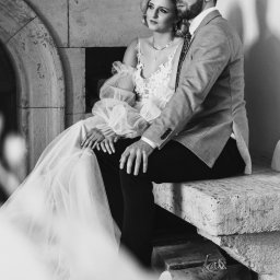 ASM PHOTOGRAPHY - Fotograf - Czarno-białe zdjęcie pary narzeczeńskiej siedzącej na kamiennej ławce przy kominku. Kobieta w sukni ślubnej z bufiastymi rękawami, mężczyzna w garniturze. Romantyczna sesja.