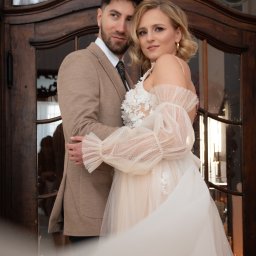 ASM PHOTOGRAPHY - Fotograf - Para narzeczeńska w objęciach na tle drewnianych drzwi z szybami. Ona w sukni ślubnej z bufiastymi rękawami, on w beżowym garniturze. Ujęcie z dołu, delikatny ruch sukni.