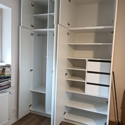 GaMeb - Biała szafa wnękowa z otwartymi drzwiami, widoczne półki i szuflady. Na podłodze panele, obok okno z książkami. Minimalistyczny design, jasne wnętrze.