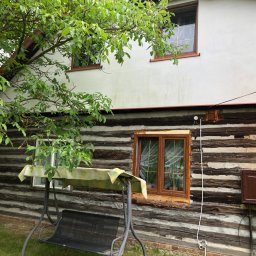 Green Watt Solar Technologies Sp. z o.o. - Drewniany dom z oknami w brązowych ramach, widoczna skrzynka elektryczna na elewacji i huśtawka ogrodowa. Częściowo otynkowana ściana i drzewo z zielonymi liśćmi.