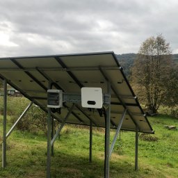 Green Watt Solar Technologies Sp. z o.o. - Instalacja fotowoltaiczna z widocznymi panelami, inwerterem Sofar i skrzynką elektryczną na tle zielonej trawy i drzew. Widok od spodu, pochmurne niebo.