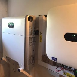 Green Watt Solar Technologies Sp. z o.o. - Biały magazyn energii Huawei zamontowany na ścianie, widoczne kable i ostrzeżenia. Element instalacji fotowoltaicznej w pomieszczeniu.