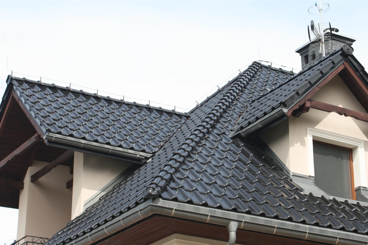 Czarny, błyszczący dach z dachówki ceramicznej na domu jednorodzinnym, widoczne orynnowanie i komin z anteną.
