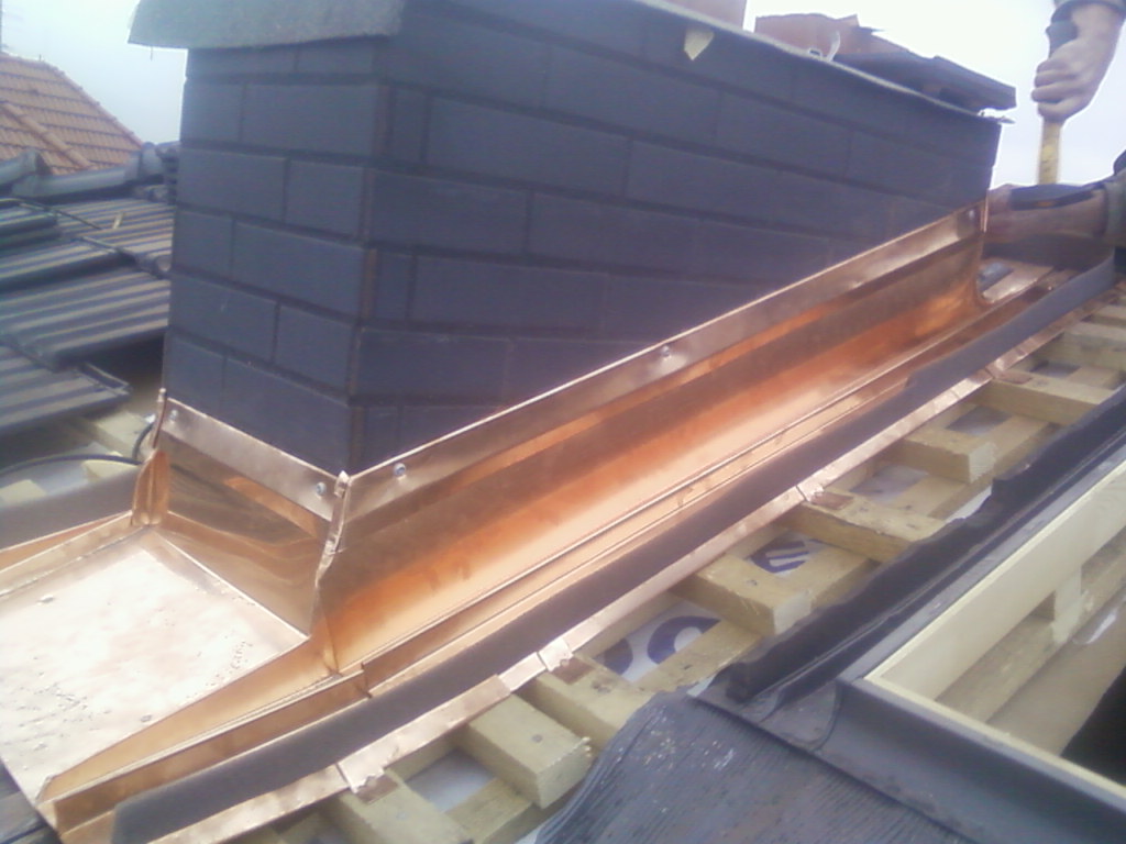 Miedziana obróbka komina z cegły klinkierowej na dachu, widoczne drewniane elementy konstrukcji dachu oraz fragment dłoni z młotkiem.