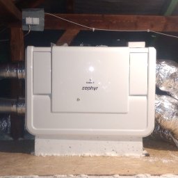 Półchłopek Instalacje sp. z o.o. - Biała centrala rekuperacyjna Zephyr w przestrzeni poddasza, z widocznymi przewodami wentylacyjnymi w izolacji aluminiowej. Drewniana konstrukcja dachu w tle.