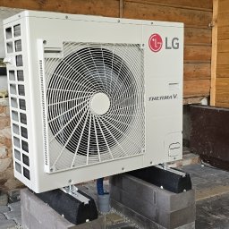 Półchłopek Instalacje sp. z o.o. - Biała pompa ciepła LG Therma V zamontowana na betonowych blokach przed drewnianym budynkiem. Widoczne elementy instalacji i otoczenie.