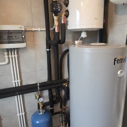 Półchłopek Instalacje sp. z o.o. - Zmontowana instalacja grzewcza z dwoma zbiornikami (TESY i Ferroli), rurami w izolacji i osprzętem. Widoczna skrzynka elektryczna i manometr. Szare płytki na ścianach i podłodze.