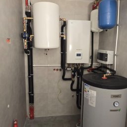 Półchłopek Instalacje sp. z o.o. - Zestaw urządzeń grzewczych: biały bojler, piec gazowy LG, zbiornik buforowy i szary zasobnik Vosti. Czarna izolacja na rurach. Szare płytki na ścianach i podłodze.