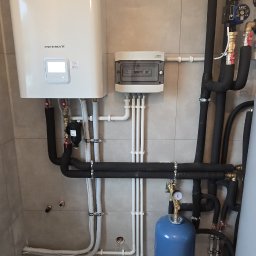 Półchłopek Instalacje sp. z o.o. - Biała pompa ciepła LG Therma V z czarnymi rurami w izolacji, niebieski zbiornik wyrównawczy i szara płytka ścienna. Widoczne liczne rury i zawory.