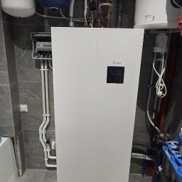 Półchłopek Instalacje sp. z o.o. - Biała pompa ciepła Midea Thermal w szarej łazience, z widocznymi rurami i zbiornikami. Nowoczesny system grzewczy w minimalistycznym wnętrzu.