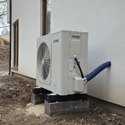 Półchłopek Instalacje sp. z o.o. - Biała pompa ciepła York zamontowana na betonowych blokach przy ścianie budynku z niebieskim przewodem. Widoczne drzewa w tle.