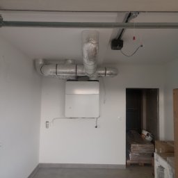 Półchłopek Instalacje sp. z o.o. - Biała centrala rekuperacyjna na ścianie, z podłączonymi, zaizolowanymi przewodami wentylacyjnymi. Widoczne elementy instalacji pod sufitem i fragment pomieszczenia.