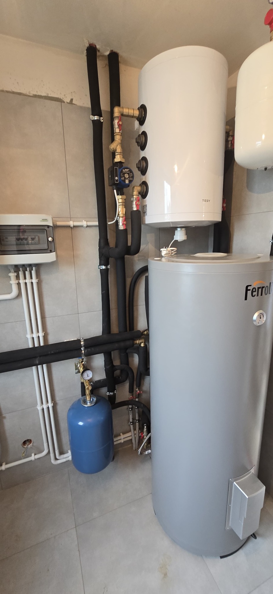 Zmontowana instalacja grzewcza z dwoma zbiornikami (TESY i Ferroli), rurami w izolacji i osprzętem. Widoczna skrzynka elektryczna i manometr. Szare płytki na ścianach i podłodze.