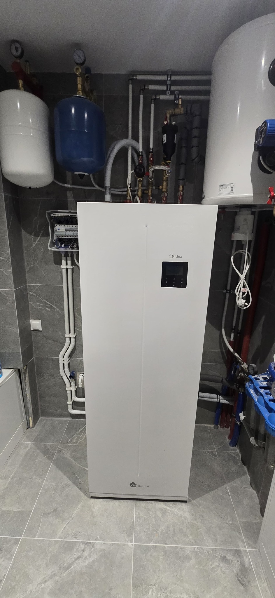 Biała pompa ciepła Midea Thermal w szarej łazience, z widocznymi rurami i zbiornikami. Nowoczesny system grzewczy w minimalistycznym wnętrzu.