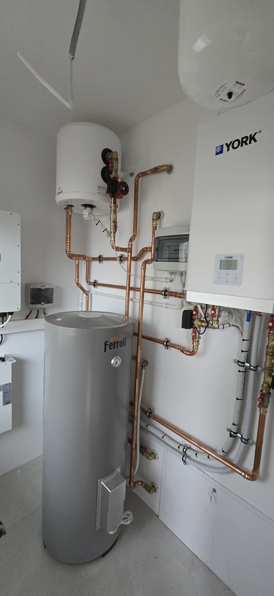 Kotłownia z piecem York, zasobnikiem Ferroli i miedzianą instalacją. Widoczne rury, zawory i elementy sterujące. Białe ściany i podłoga. Nowoczesny system grzewczy.