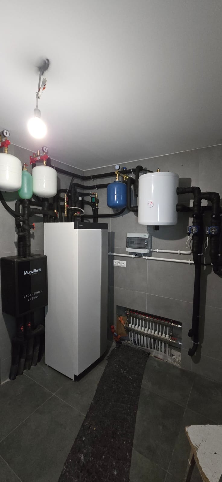 Złożona instalacja grzewcza z kotłem Muovitech, zbiornikami i rozdzielaczem podłogówki w szarej kotłowni. Widoczne liczne rury i zawory.