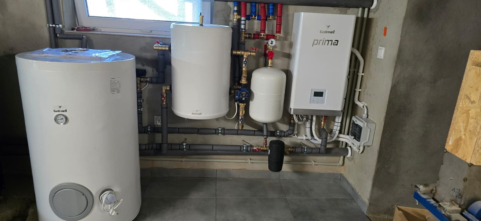 Białe zbiorniki Galmet i instalacja grzewcza z rurami w szarym pomieszczeniu z oknem. Widoczne zawory, manometry i inne elementy instalacji. Szare płytki na podłodze.