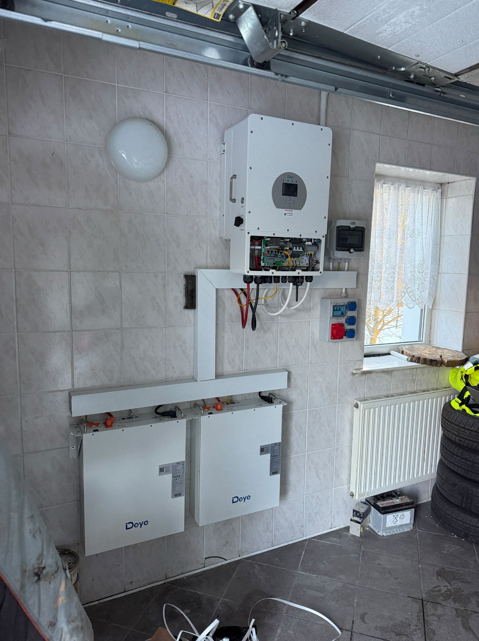 Biały inwerter i akumulatory Deye zamontowane na ścianie z kafelków w garażu, widoczne okablowanie i elementy instalacji elektrycznej. W tle okno i grzejnik.