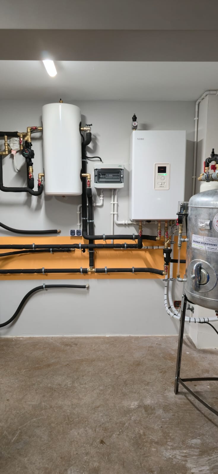 Zestaw urządzeń grzewczych Nibe z zbiornikiem hydroforowym i rozbudowaną instalacją rur miedzianych i czarnych, zamontowany na ścianie w pomieszczeniu technicznym.