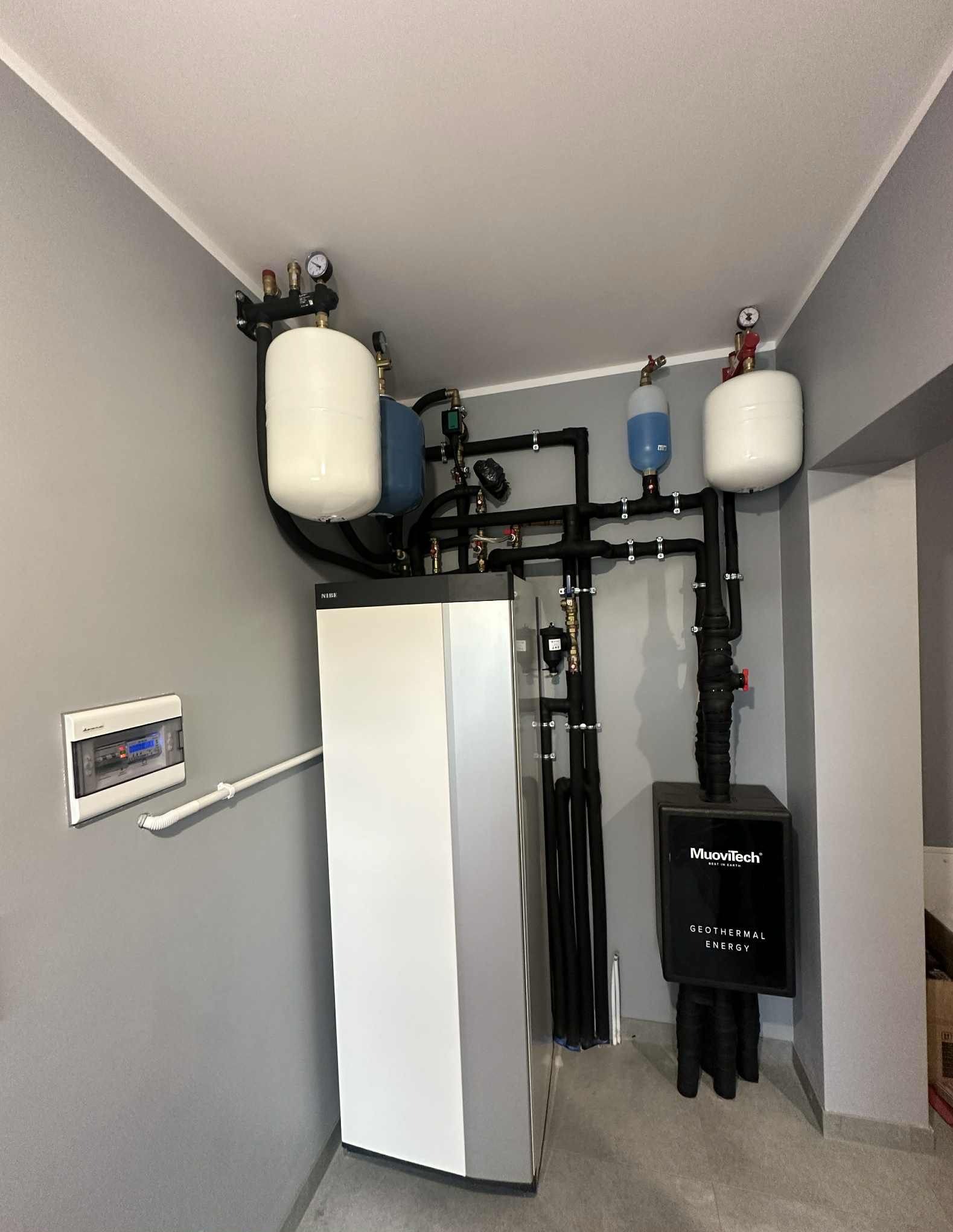 Zmontowana instalacja pompy ciepła Muovitech z zbiornikami buforowymi i czarnymi rurami w szarej kotłowni. Widoczny panel sterowania na ścianie.