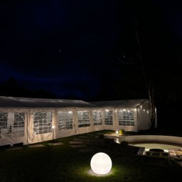 All4events - Oświetlony, biały namiot eventowy z przezroczystymi ścianami, ustawiony na trawniku nocą. Dekoracyjne oświetlenie zewnętrzne, ścieżka z kamieni i altana w tle.