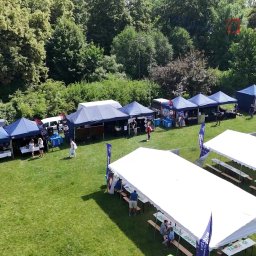 All4events - Widok z góry na rozstawione białe i niebieskie namioty na zielonej trawie podczas plenerowej imprezy. W tle drzewa i krzewy, a także food trucki.