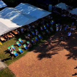 All4events - Z lotu ptaka: białe namioty eventowe z ławami i rzędy zielono-niebieskich leżaków na trawie. Ludzie odpoczywają na słonecznym placu z bruku.