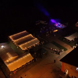 All4events - Nocne wydarzenie plenerowe z lotu ptaka: oświetlone namioty eventowe, scena z fioletowym światłem i bawiący się goście.