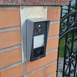 Go Sat Cam - Zamontowany wideodomofon HiLook na ceglanym słupku ogrodzenia z granitowym wykończeniem. Widoczna brama z kutego żelaza i zieleń w tle. Estetyczny montaż.