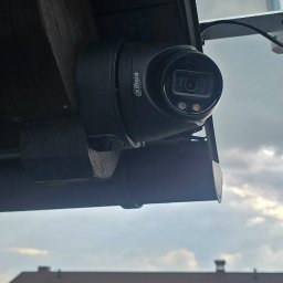Go Sat Cam - Kamera monitoringu Dahua zamontowana pod drewnianym okapem, skierowana na budynek gospodarczy z żółtą elewacją i brązowym dachem. Widoczne pochmurne niebo.