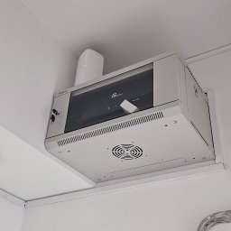 Go Sat Cam - Zamontowana pod sufitem szafa rack z routerem. Widoczne kable i wentylator. Białe ściany i sufit. Fragment drabiny malarskiej w prawym dolnym rogu.
