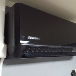 Klima Instalator - Czarny klimatyzator Rotenso z białym przewodem na szarej ścianie. Widoczny wyświetlacz temperatury i detale wykończenia. Ujęcie z dołu.