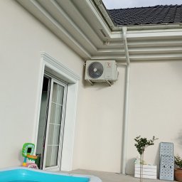 Klima Instalator - Zamontowany klimatyzator Rotenso na elewacji domu, widoczny z perspektywy podwórka z basenem dla dzieci. Biała ściana, rynna i fragment dachu. Pochmurne niebo w tle.