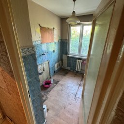 Śląski Multiserwis - Zdemontowana kuchnia z częściowo usuniętymi płytkami, widoczne ślady po instalacjach. W tle okno z widokiem na zieleń. Ujęcie przez otwarte drzwi.