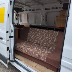 Usługi transportowe Bartłomiej Borowczyk - Biały bus z otwartymi drzwiami bocznymi, w środku brązowa sofa z liściastym wzorem, drewniany stolik i kartony. Podłoga przestrzeni ładunkowej zabezpieczona tekturą.