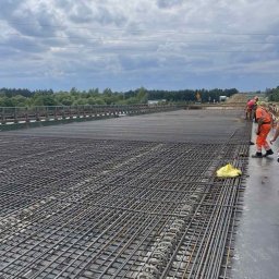 zbrojenia.eu - Rozległe zbrojenie mostu z widocznymi pracownikami w pomarańczowych kombinezonach i zielenią w tle pod pochmurnym niebem.