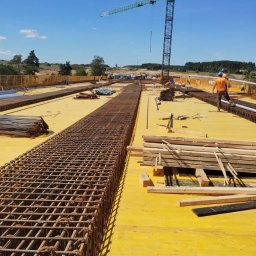 zbrojenia.eu - Zbrojenie fundamentów na żółtej płycie budowlanej, widok z góry. Na drugim planie dźwig budowlany i pracownicy w pomarańczowych koszulkach.