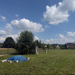 Ogrody kompleksowe usługi - Duże, świeżo skoszone pole trawiaste w Gorlicach, z widocznymi pokosami, pod błękitnym niebem z białymi chmurami. W tle domy i drzewa, a na pierwszym planie niebieska plandeka i betonowe bloki.