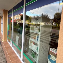 EcoPure Cleaning Solutions - Witryna sklepowa z białymi ramami i niebiesko-zielonymi pasami, odbijająca ulicę i niebo. Wewnątrz widoczne produkty Ziaja na półkach.