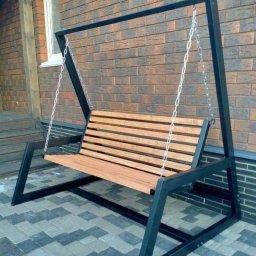 patspaw - Metalowa huśtawka ogrodowa z drewnianym siedziskiem, zawieszona na łańcuchach, na tle ceglanej ściany i kostki brukowej. Minimalistyczny design, czarna rama.