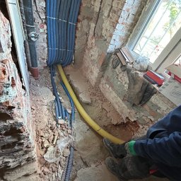 Belweder Energy - Niebieskie i żółte rury karbowane prowadzone w bruzdach w ścianie z czerwonej cegły, widoczne fragmenty instalacji i okno w tle. Prace instalacyjne w trakcie remontu.