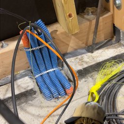 Belweder Energy - Niebieskie rury kablowe przymocowane do ściany, z czarnymi i pomarańczowymi kablami w tle. Kot w czapce w pierwszym planie dodaje unikalnego charakteru.