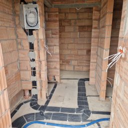 Belweder Energy - Ułożone czarne peszle instalacji elektrycznej na betonowej posadzce i w ścianach z pustaków ceramicznych, z widoczną otwartą rozdzielnicą i niebieskim przewodem.