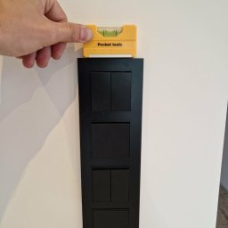 Belweder Energy - Dłoń poziomuje czarny, nowoczesny panel z czterema włącznikami na jasnej ścianie, używając żółtej poziomicy 'Pocket tools'.