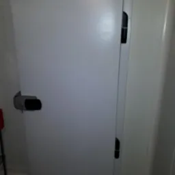 Białe drzwi chłodnicze z zamkiem i kluczem, widoczne w pomieszczeniu z panelami ściennymi.